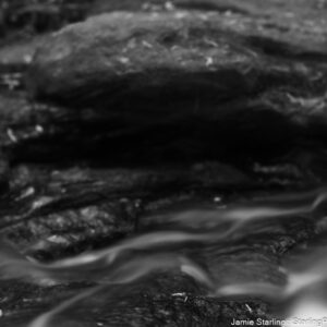 A black and white photograph showcasing the gentle flow of water over the textured surface of rocks, where the interplay of light and shadow captures the serene movement in a timeless frame.