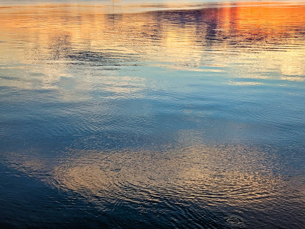 A serene sunset over water with ripples reflecting a fusion of fiery orange and calming blue, symbolizing life's joy and serenity.