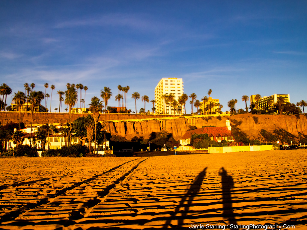 A beautiful sunset casting long shadows over golden sands, with palm trees gently swaying and buildings in the distance, evoking a sense of peace and inspiration for creative expression.