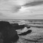 A captivating black and white photograph of a cliff overlooking the ocean with waves crashing against the rocks and a sunlit, cloudy sky, symbolizing a journey of self-discovery and personal growth.