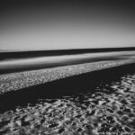 A black-and-white photo of a beach at dusk with long shadows cast by the dunes and smooth, flowing waves in the background, symbolizing the balance of light and shadow in life.