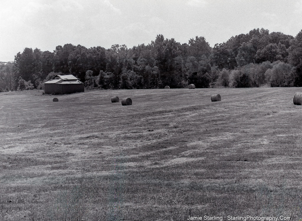 A black and white image of a rural field with hay bales and a weathered barn, symbolizing patience, hard work, and the enduring rhythms of nature.