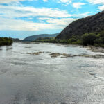 A calm river flows between green hills under a partly cloudy sky, symbolizing the harmony between going with the flow and taking necessary actions in life.