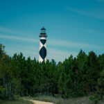 A black-and-white lighthouse rises above green trees with a winding path leading toward it, symbolizing guidance and the journey of self-discovery—a striking visual metaphor in photography.