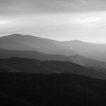 Black-and-white photograph of layered mountain ridges with light breaking through clouds, symbolizing the power of perspective and the depth of life’s layers.