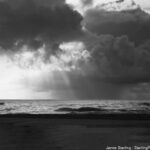 Dark clouds over the ocean with rays of light breaking through, symbolizing moments of clarity piercing through illusions.