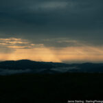 A peaceful mountain sunset with rays of light breaking through the clouds, symbolizing the balance between light and darkness and inviting a deeper connection with nature.