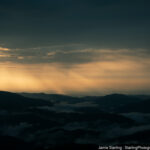 Sunlight breaking through clouds over misty mountain ridges, symbolizing the journey from blindness to insight and uncovering hidden truths.