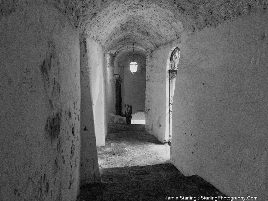 A quiet alleyway in black and white with soft light from a single lantern, highlighting rough-textured walls and shadows, inviting the viewer to pause and reflect on the beauty of simple spaces.