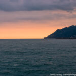 A serene ocean view at sunset with a soft orange glow on the horizon and distant mountains, inviting you to slow down and appreciate the moment.