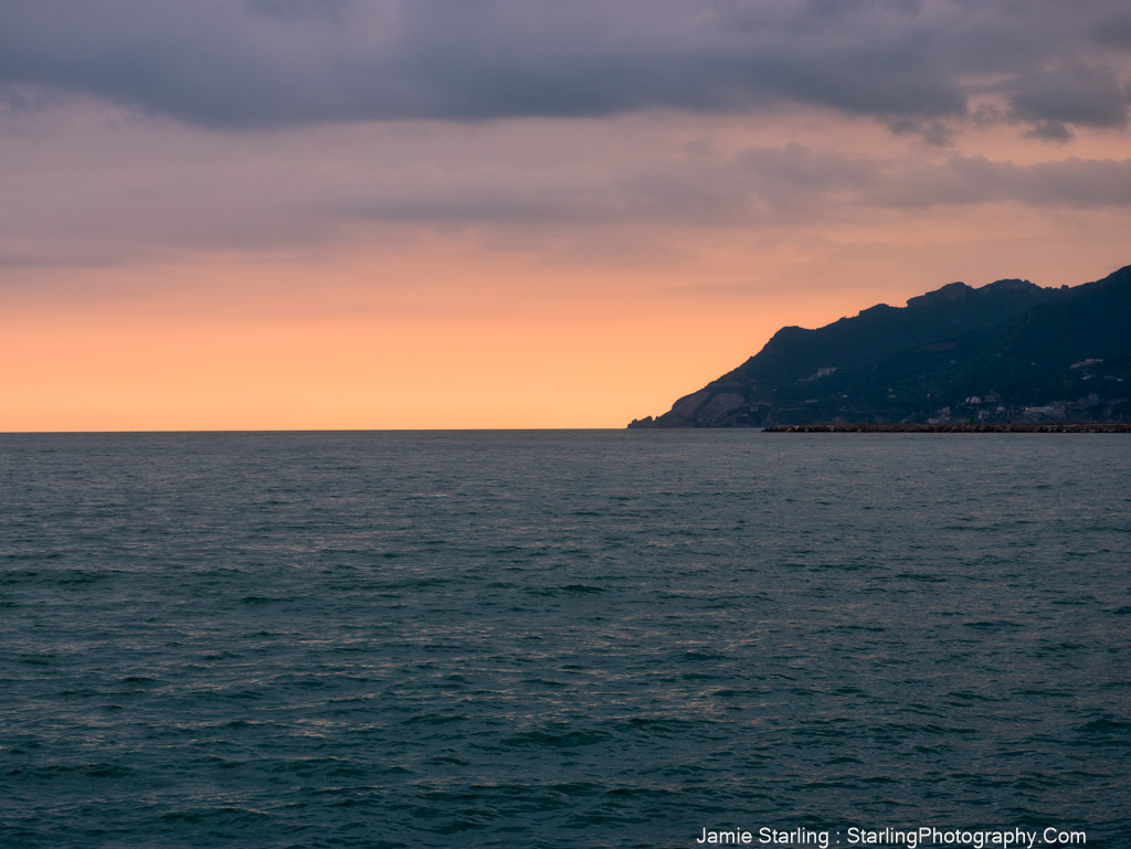 A serene ocean view at sunset with a soft orange glow on the horizon and distant mountains, inviting you to slow down and appreciate the moment.