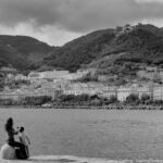 A peaceful black-and-white photo of a mother brushing her child’s hair by the water, with a hillside town and mountains in the background, capturing a moment of care and connection.