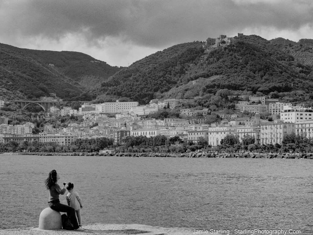 A peaceful black-and-white photo of a mother brushing her child’s hair by the water, with a hillside town and mountains in the background, capturing a moment of care and connection.